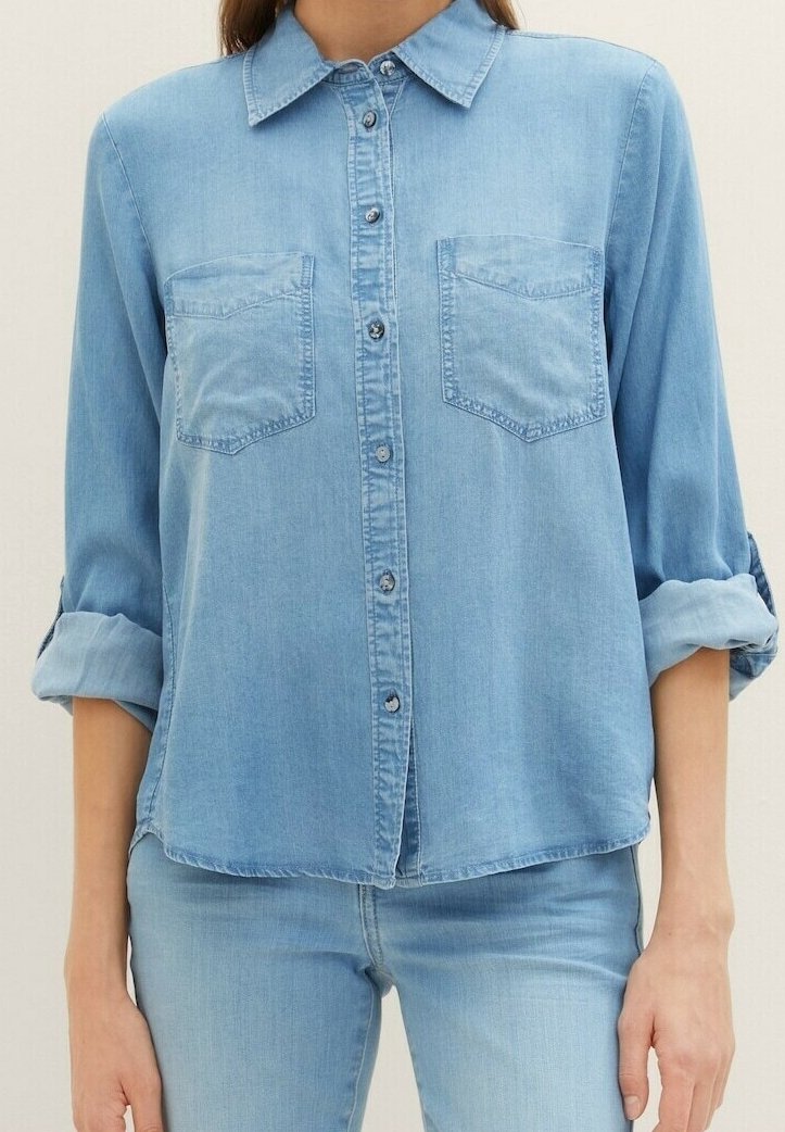 Femme portant une chemise en denim bleu clair à boutons avec deux poches poitrine et manches retroussées, associée à un jean bleu clair.
