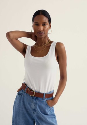 Femme aux cheveux foncés portant un débardeur blanc, un jean bleu avec une ceinture marron, des boucles d'oreilles créoles en or et des colliers en or superposés, posant avec une main derrière la tête.