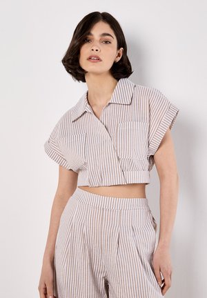 Blusa corta de manga corta con un patrón a rayas blanco y beige claro, cuello abotonado y dos bolsillos en el pecho.