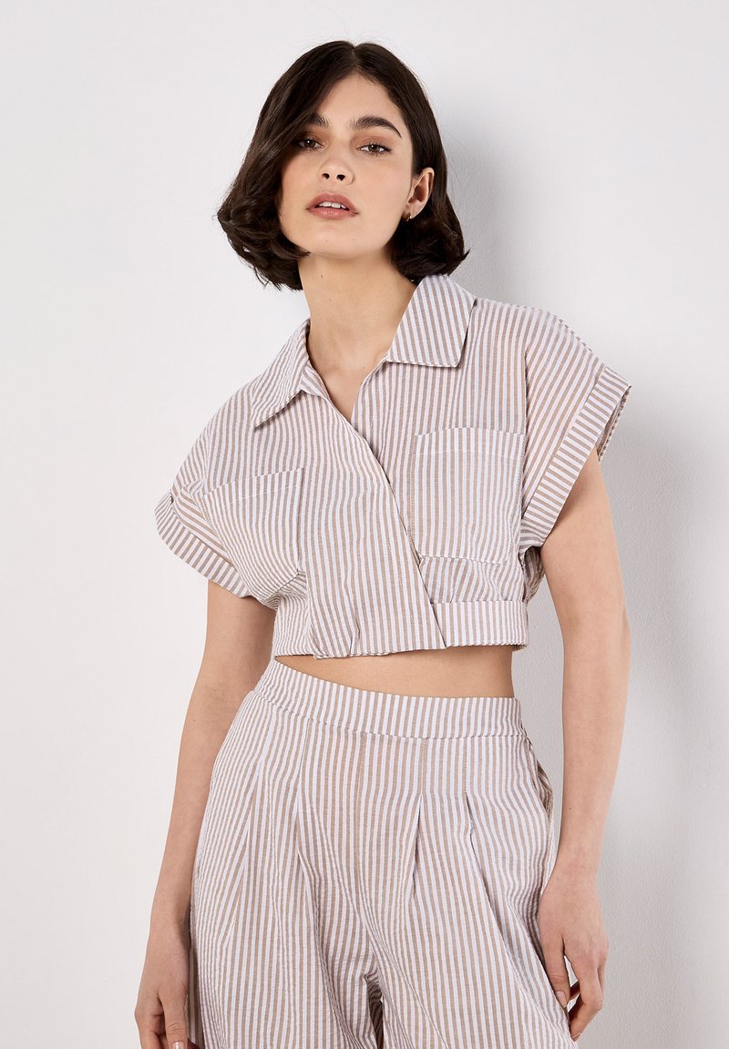 Blusa corta de manga corta con un patrón a rayas blanco y beige claro, cuello abotonado y dos bolsillos en el pecho.