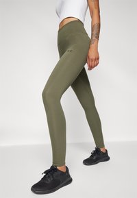 Sieviete, kas valkā olīvu zaļas augstā vidukļa leggingas un melnas sporta kurpes, stāv ar vienu kāju nedaudz saliektu un redzamu tetovētu roku.