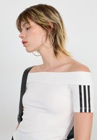 Balta off-shoulder palaidinė su juodomis akcentinėmis juostelėmis ant rankovių, lygi tekstūra. Modelis dėvi dideles auskarus ir laiko juodą rankinę su dirželiu.
