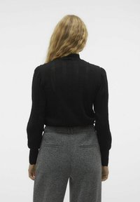 Zwarte gebreide trui met een tekstuurd kabelpatroon, hoge halslijn en lange mouwen; gedragen met grijze gebreide broek.