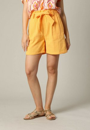 Shorts en tissu texturé orange avec une large ceinture et un détail à nouer, associés à des sandales à lanières croisées beiges et un haut à motifs.