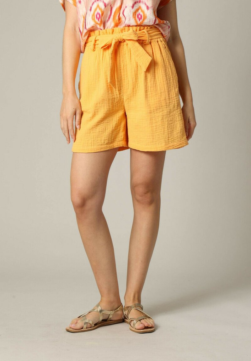 Shorts en tissu texturé orange avec une large ceinture et un détail à nouer, associés à des sandales à lanières croisées beiges et un haut à motifs.