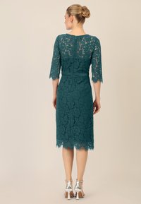 Abito in pizzo teal con maniche a tre quarti, orlo a onde e vita aderente. Presenta una chiusura con zip posteriore e sandali argento.
