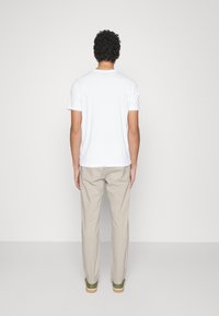 Man med lockigt hår iklädd en vit t-shirt med korta ärmar, beige byxor och gröna sneakers står vänd mot en enkel ljus vägg.