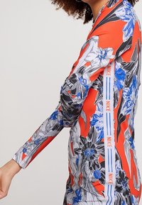 Donna che indossa una maglietta Nike a maniche lunghe con un motivo floreale nei colori rosso, bianco, blu e nero, con strisce del logo Nike lungo il lato.