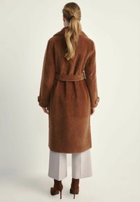 Kobieta z blond włosami w kucyku, ubrana w brązowy sweter wełniany z paskiem, białe spodnie z szerokimi nogawkami oraz brązowe szpilki, odwrócona tyłem.