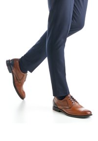 Brauner Lederschuh für Damen mit rundem Zehenbereich, Schnürdesign und dezenten Ziernähten; getragen zu marineblauen Hosen und dunklen Socken.