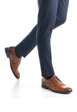 Brauner Lederschuh für Damen mit rundem Zehenbereich, Schnürdesign und dezenten Ziernähten; getragen zu marineblauen Hosen und dunklen Socken.