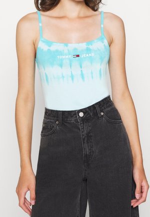 Kobieta nosząca niebiesko-biały top na cienkich ramiączkach z logo „Tommy Jeans”, w zestawieniu z czarnymi jeansami z wysokim stanem.