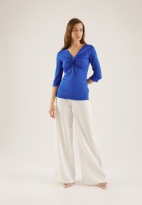 Blue, long-sleeve top with a gathered twist detail at the front, paired with white wide-leg trousers and nude pointed-toe heels.