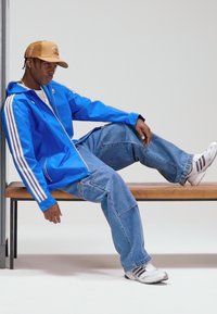 Blå zip-up jacka med vita ränder, lösa jeans i denim och sportiga vita sneakers. Modellen sitter på en träbänk i en neutral miljö.