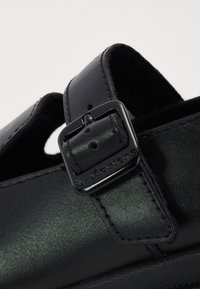 Black leather shoe with a single strap featuring a metal buckle. Smooth texture and subtle stitching detail. Brand logo visible on the strap.