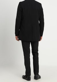 Giacca nera con una vestibilità su misura, texture liscia e design della schiena dritto, abbinata a pantaloni neri coordinati e scarpe eleganti.