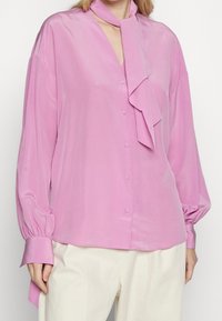 Blusa rosa chiaro con maniche lunghe e gonfie, scollatura a V e dettaglio di una sciarpa annodata al colletto. Tessuto liscio con bottoni sulla parte anteriore.