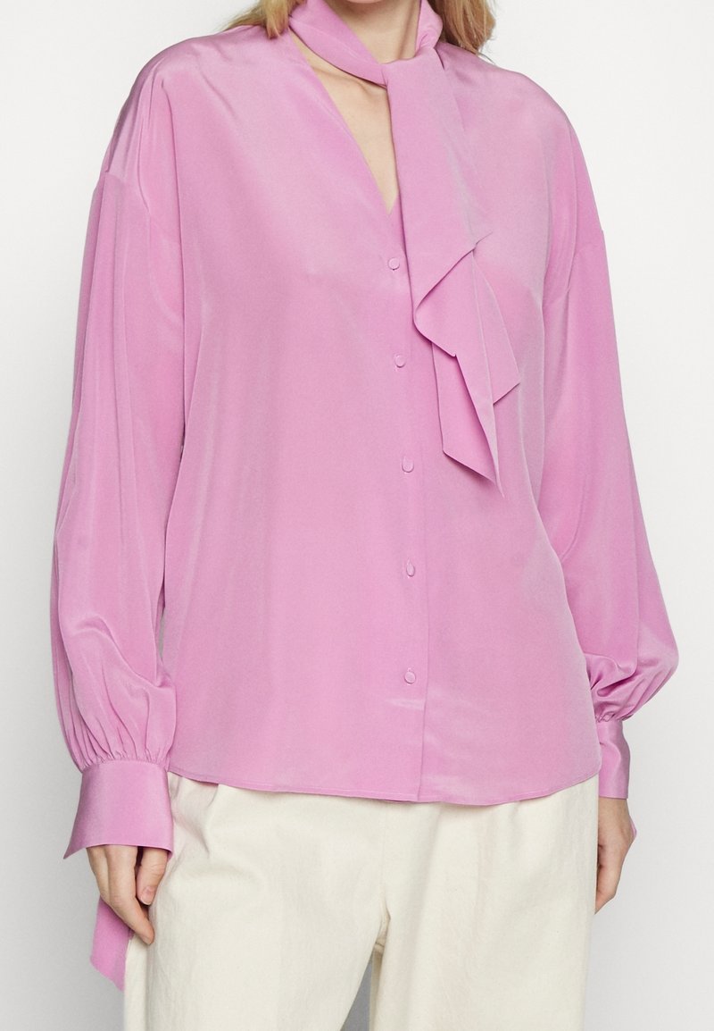 Blusa rosa chiaro con maniche lunghe e gonfie, scollatura a V e dettaglio di una sciarpa annodata al colletto. Tessuto liscio con bottoni sulla parte anteriore.