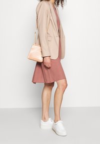 Blazer beige chiaro su un vestito rosa polveroso, abbinato a sneakers bianche e una piccola borsa pesca texture. Colori neutri e linee pulite.