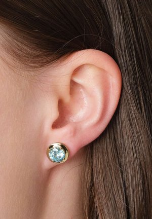 Nahaufnahme eines Ohrs mit dunklem Haar, das einen goldenen Steckerohrring mit einem runden, hellblauen Edelstein trägt.