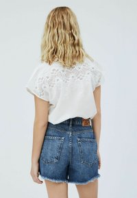 Bílý top s krátkým rukávem a krajkovými detaily na zádech, doplněný modrými džínovými šortkami s roztrhaným lemem a dvěma zadními kapsami.