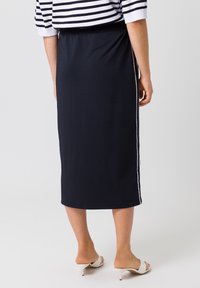 Jupe midi noire en tissu doux, avec une texture lisse et des rayures latérales blanches. Associée à des chaussures ouvertes de couleur claire.