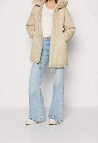 Abrigo acolchado beige con capucha, con cierre de cremallera y cordón en la parte inferior, combinado con jeans acampanados azul claro y zapatillas crema.