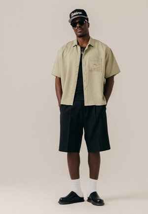 Homme portant une chemise beige à manches courtes boutonnée, un short noir, des chaussettes blanches, des chaussures noires sans lacets, des lunettes de soleil noires et une casquette noire des Raiders.