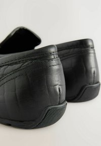 Chaussures noires en cuir à enfiler avec une finition texturée, un talon arrondi et une semelle en caoutchouc. Présente des coutures contrastées et un design supérieur flexible.