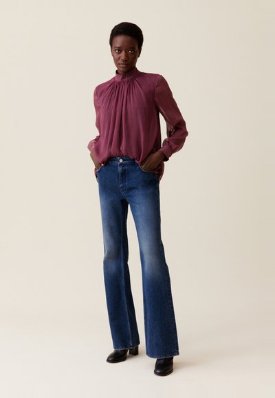 Mujer de pie con las manos en los bolsillos, vestida con una blusa burdeos de cuello alto y vaqueros de pierna ancha azules, con botas negras hasta el tobillo.