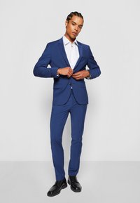 Marineblauw pak met een gestructureerd colbert en slim-fit broeken, gecombineerd met een wit, gestreept overhemd en zwarte veterschoenen.