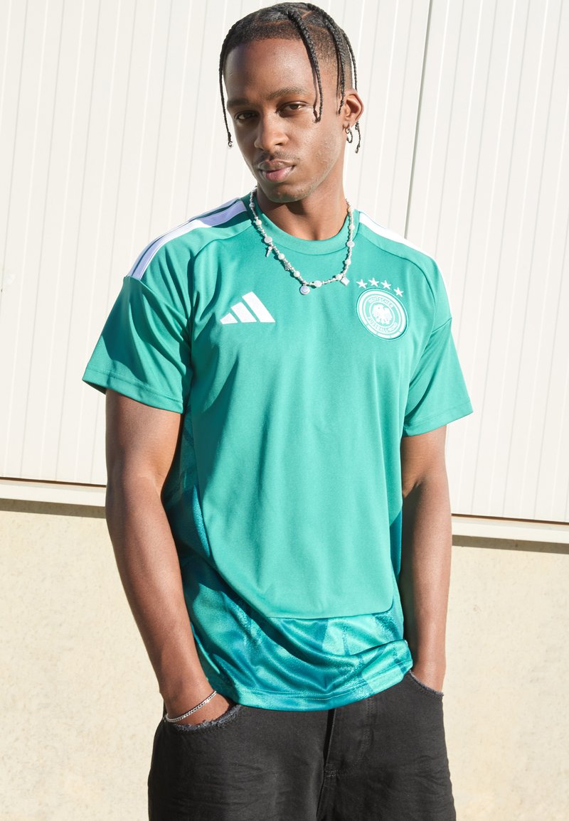 Camisola atlética verde com detalhes brancos, apresentando um gola redonda, mangas curtas e emblema da equipa. Usada com calções pretos e acessórios.