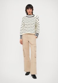 Dryžuotas megztinis grietinėlės ir jūrinės spalvos su apvaliu iškirpimu ir mygtukų detale. Šviesiai rudos spalvos kargo kelnės su kišenėmis ir platėjančiomis kojomis. Juodi aulinukai iki kulkšnies.