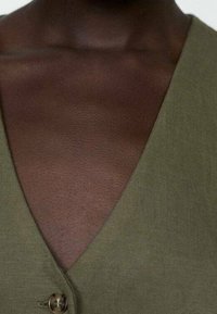 Olive green top z dekoltem w kształcie litery V, wykonany z teksturowanej tkaniny, z detalami w postaci guzika na środku przodu. Zbliżenie na dekolt i obszar klatki piersiowej.