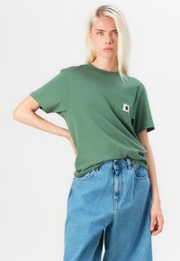 Olivenfarget bomulls t-skjorte med rund hals og korte ermer, med en liten lomme med logo detalj. Kombinert med lyseblå denim jeans.