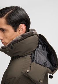 Olijfgroene gewatteerde jas met een faux fur kraag, voorzien van een rits en een zwarte binnenvoering. Details zijn onder andere drukknopen en trekkoorden.