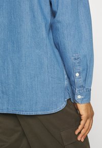 Chemise en denim bleu clair avec un poignet boutonné et un ourlet arrondi. Présente une texture lisse et des détails de couture subtils. Pantalon cargo marron visible.