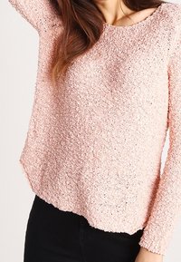 Femme portant un pull à manches longues texturé rose clair avec un pantalon noir, bras levé, montrant le tissu doux et confortable du pull.
