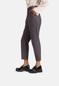 Pantaloni grigi su misura con una vestibilità slim, dotati di tasche laterali e pieghe ben definite, abbinati a mocassini neri in pelle lucida con dettagli testurizzati.