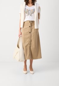 Béžová bavlněná sukně s předními knoflíky, bílý tričko se zdobným potiskem a krémový kardigan. Modelka drží světlou tašku a má na sobě béžové lodičky.