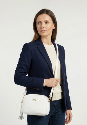 Žena v námořnicky modrém blejzru a krémovém svetru drží bílou kabelku s přezkou a zlatými detaily, dívá se stranou na jednoduchém pozadí.