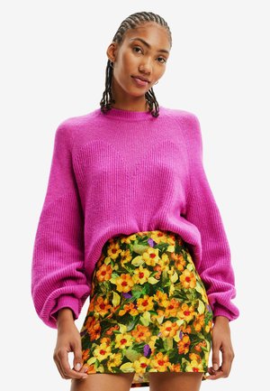 Woman with braided hair wearing a bright pink knit sweater and a vibrant floral mini skirt with orange and yellow flowers.