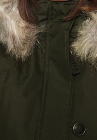 Parka verde oscuro con cuello de piel sintética, gran botón delantero y textura sutil. Cuenta con cremallera y lazos de tela en el cuello.