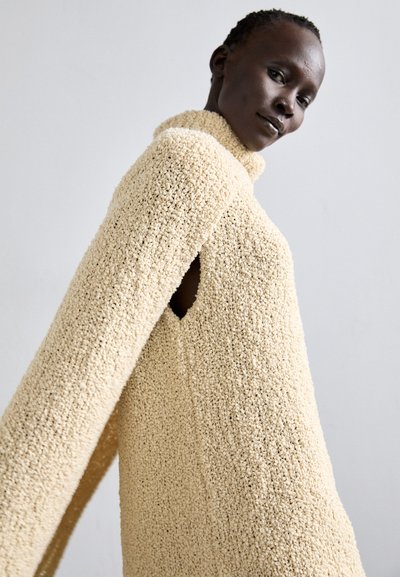 Cream-colored sweater dress with a textured, fuzzy fabric. Features a high neck and open armholes, providing a draped silhouette.