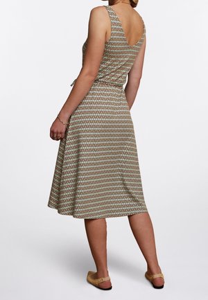 Femme portant une robe sans manches à motifs avec un dos en V et des chaussures plates beiges, debout devant un fond blanc uni.