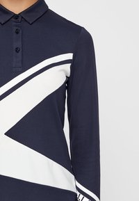 Mörkblå pikétröja med långärmad och vitt geometriskt mönster. Har klassisk krage, treknappslinning och strukturerat tyg.