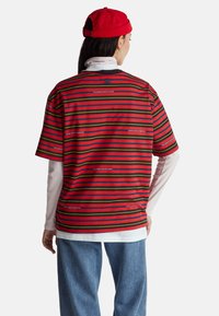 Camisa de manga corta a rayas rojas y verdes con acentos negros, usada sobre una camiseta blanca de manga larga, combinada con vaqueros azules holgados.