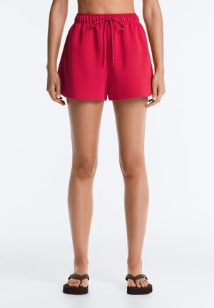 Nő élénkvörös, zsinóros rövidnadrágot és fekete platform papucsot visel, fehér háttér előtt áll.