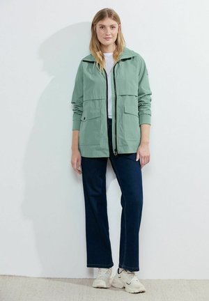Veste zippée vert clair avec deux poches avant, portée sur un haut blanc. Associée à un jean bleu foncé et des baskets claires.