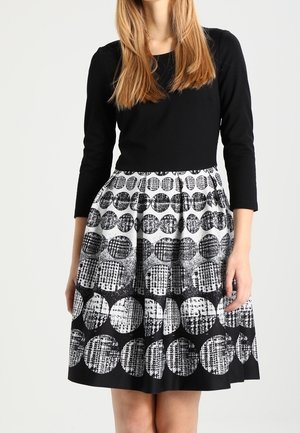 Femme portant un haut noir à manches longues et une jupe longueur genou avec un motif circulaire texturé noir et blanc.